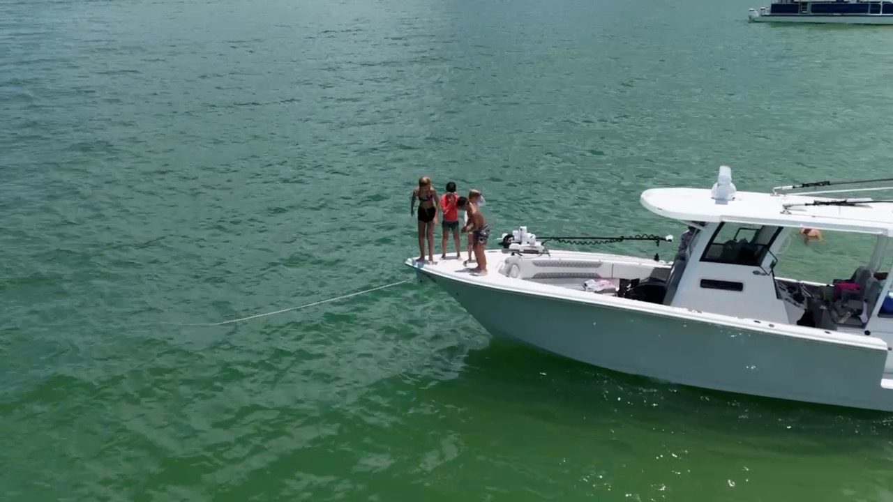 Aerial view of a white center-console boat anchored in green water with kids standing on the bow